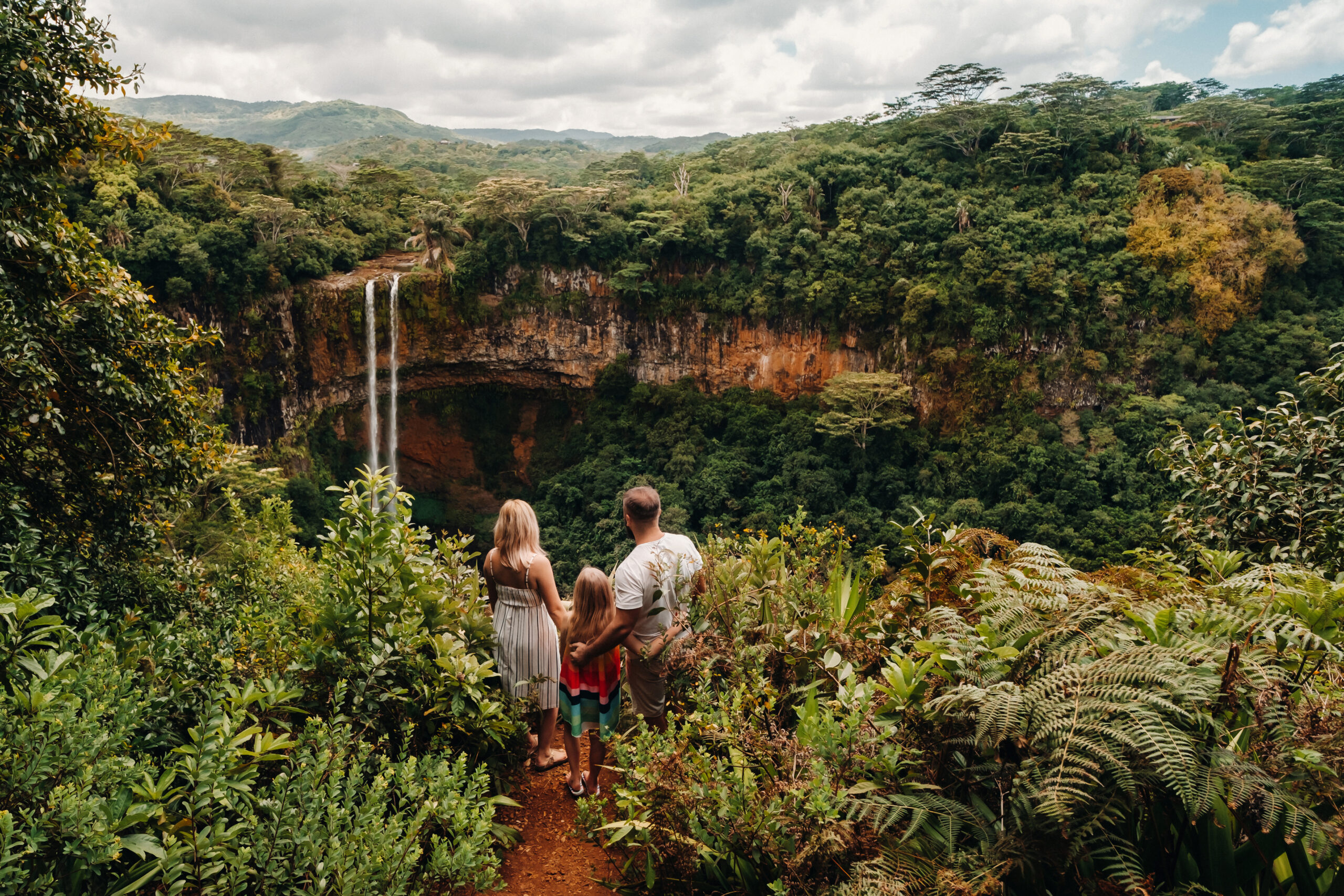 a family, a man, a woman and a daughter, stand on the edge of a cliff near a waterfall in Chamarel Park on the island of Mauritius.A couple with their daughter in the jungle of the island of Mauritius looks at a large waterfall falling down.